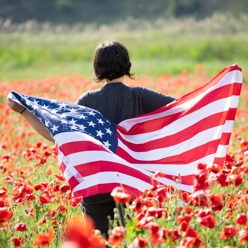 Elevate Your Memorial Day Celebrations with a Natural Hemp Treat - Modern CBD & Wellness - Modern CBD & Wellness Person viewed from behind stands in a field of red flowers, holding a large American flag draped around their shoulders.