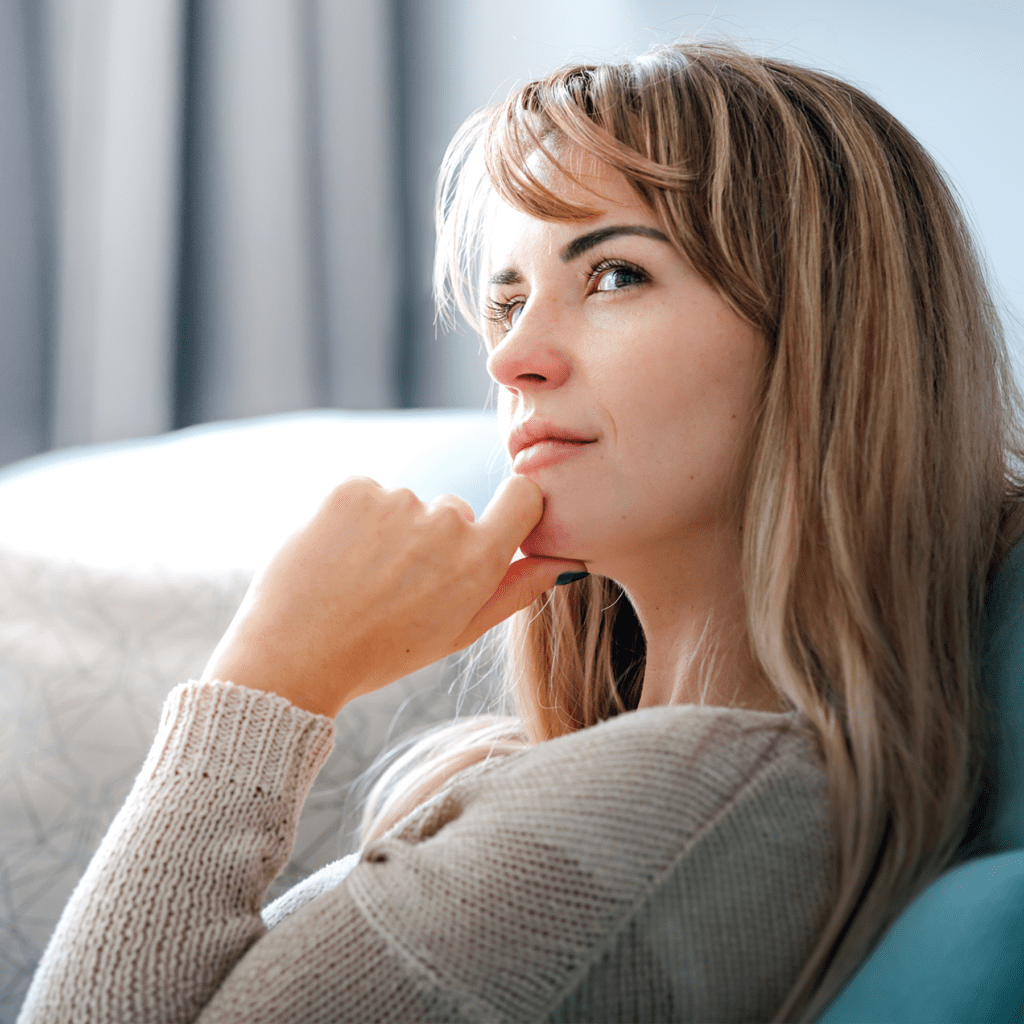 Close-up of a woman indoors, resting her chin on her hand, with a thoughtful expression and a beige knitted sweater.